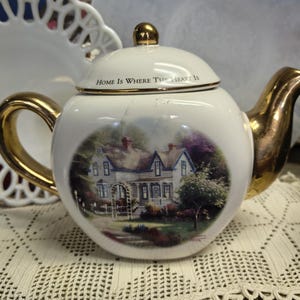 May include: A white ceramic teapot with gold accents on the handle, spout, and lid. The teapot features a scenic image of a house and the text "Home Is Where The Heart Is" on the lid. The teapot is sitting on a lace tablecloth.
