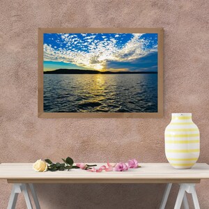 Peut inclure: Une impression de paysage encadrée rectangulaire montrant un coucher de soleil sur une étendue d'eau avec la silhouette sombre d'une terre au loin, un ciel bleu et jaune vif avec des nuages, un vase jaune et une rose crème aux pétales roses.