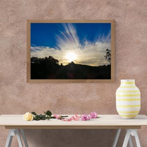 Peut inclure: Une photographie de paysage encadrée montrant un coucher de soleil sur une silhouette sombre de collines et d'arbres sous un ciel bleu et blanc avec des nuages effilés. À côté du cadre se trouve un vase en céramique rayé blanc et jaune.