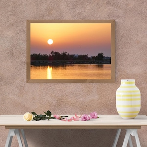 Peut inclure: Une impression d'art mural encadrée rectangulaire représentant un coucher de soleil sur un plan d'eau calme avec la silhouette d'arbres et de terre au loin. Le ciel présente un dégradé de tons orange et jaune, reflétant la lumière sur l'eau.