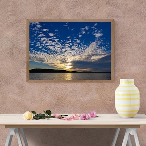 Peut inclure: Une photographie de paysage encadrée représentant un coucher de soleil sur une étendue d'eau sombre avec une chaîne de montagnes en silhouette au loin et un ciel rempli de nuages blancs épars. Un vase rayé jaune et blanc est posé sur une table.