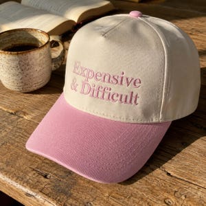 May include: A cream-colored baseball cap with a pink brim and a pink button on top. The front of the cap features the embroidered text "Expensive & Difficult" in pink lettering. The cap has a structured five-panel design with visible stitching.