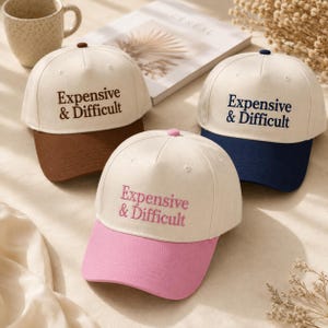 Peut inclure: Trois casquettes de baseball avec des calottes blanc cassé et des visières colorées. Chaque casquette présente le texte brodé Expensive & Difficult. Une casquette a une visière et un texte marron, une bleu marine, et une rose.