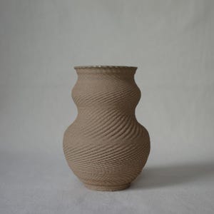 Puede incluir: Un jarrón de color beige con una forma de reloj de arena distintiva y un patrón texturizado en espiral en toda su superficie. El recipiente presenta un cuello estrecho que se ensancha hacia un cuerpo redondeado, con una base plana y borde.
