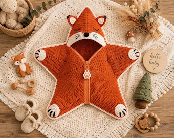 May include: A star-shaped crochet baby swaddle blanket in burnt orange with a fox hood featuring embroidered eyes, whiskers, and a black nose. The garment includes a central zipper with a white paw-shaped pull and white paw-pad accents on the tips.