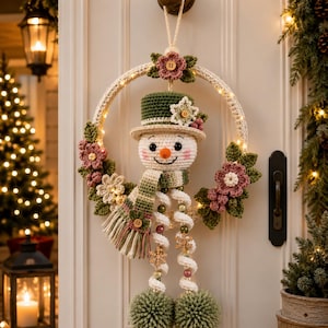 May include: A crocheted snowman wreath featuring a green top hat, a striped scarf, and dangling legs with pom-poms. The circular frame is adorned with crocheted pink flowers, green leaves, and small wooden buttons, illuminated by integrated warm white fairy lights.