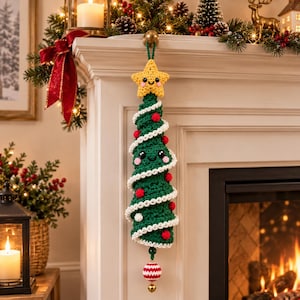 Op de afbeelding: Een gehaakte kerstboomversiering met een groen spiraalvormig lichaam, witte randen, kleine rode pompons en een lachende gele ster aan de bovenkant. Het ornament heeft een vriendelijk gezicht met zwarte ogen en roze wangen aan de onderkant.