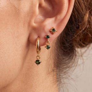 May include: A set of three gold-toned earrings featuring small green gemstones. One earring is a hoop with a dangling green stone, another is a stud with two vertical green stones, and the third is a star-shaped stud with a central green stone.