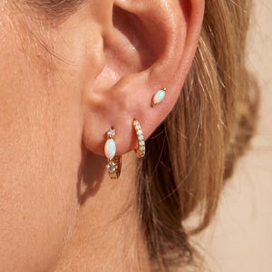 May include: A gold hoop earring featuring a marquise-cut opal and a small round clear stone, a plain gold hoop earring, and a small gold stud earring with a marquise-cut opal, all worn in a single earlobe with multiple piercings.