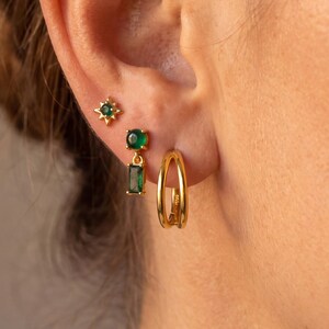 May include: A set of three gold-toned earrings worn in the earlobe and cartilage. The top earring is a small star-shaped stud with a green stone, the middle is a drop earring with a round and rectangular green stone, and the bottom is a double-band hoop.