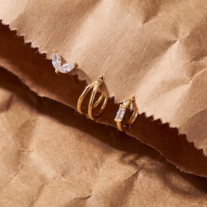 May include: Three gold-toned earrings are displayed. One earring features two marquise-cut clear stones, another is a triple-band hoop, and the third is a small hoop earring set with a single rectangular baguette-cut clear stone.