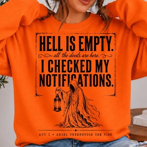 May include: A bright orange long-sleeve sweatshirt featuring black text that reads HELL IS EMPTY. all the devils are here. I CHECKED MY NOTIFICATIONS. Below the text is a black graphic illustration of a hooded figure holding a lantern.
