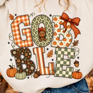 May include: A cream-colored crewneck sweatshirt featuring the text GOBBLE in large block letters. Each letter has a unique pattern including orange plaid, leopard print with a turkey, pumpkins, floral print, orange stripes, and green checkered patterns.