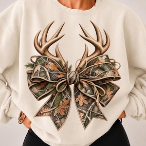 May include: A cream-colored crewneck sweatshirt featuring a large graphic print on the front. The design displays a pair of brown deer antlers positioned above a decorative bow with a camouflage pattern, accented with green pine needles and leaves.
