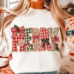 May include: A white long-sleeve sweatshirt featuring the word MERRY in large, decorative letters. The letters are patterned with red and green plaid, adorned with festive bows, holly leaves, berries, and a small heart and tree tag.