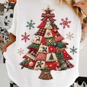 Può includere: Una t-shirt bianca con una grande grafica di un albero di Natale composto da vari ritagli di tessuto rosso e verde con motivi scozzesi, vichy e a tinta unita. L'albero è decorato con piccoli fiocchi, ornamenti e fiocchi di neve.