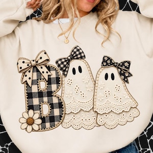 Könnte beinhalten: Ein cremefarbenes Sweatshirt mit Rundhalsausschnitt und einem Aufdruck des Wortes BOO. Der Buchstabe B ist schwarz-weiß kariert mit einem Gänseblümchen und einer gepunkteten Schleife. Zwei weiße Geister mit Spitzendetails und Schleifen stehen daneben.