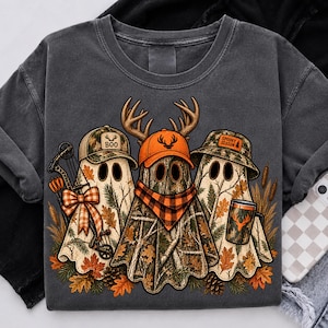 May include: A charcoal gray t-shirt featuring a graphic of three ghosts wearing camouflage hats. The left ghost holds a bow with an orange plaid bow, the center ghost wears an orange cap and plaid scarf, and the right ghost holds a mug labeled SPOOKY SEASON.
