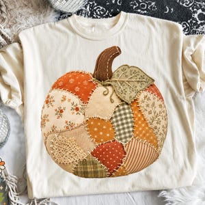 May include: A cream-coloured t-shirt featuring a large, central graphic of a pumpkin constructed from various fabric patches. The pumpkin design includes diverse patterns like floral prints, gingham, polka dots, and lace, all joined by visible stitching.