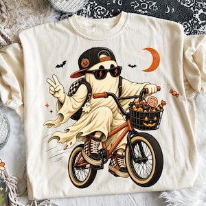 May include: A cream-colored t-shirt featuring a graphic of a ghost wearing a black baseball cap with a pumpkin icon, sunglasses with lightning bolts, and checkered sneakers. The ghost is riding an orange bicycle with a jack-o'-lantern basket.