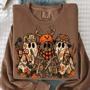 May include: A brown crewneck sweatshirt featuring a graphic of three ghosts wearing camouflage hats. The ghosts are decorated with autumn leaves, a bow, a compound bow, a plaid scarf, and a mug. The text reads BOO and SPOOKY SEASON.