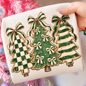 Può includere: Una t-shirt bianca con una stampa grafica di tre alberi di Natale stilizzati. L'albero a sinistra ha un motivo a scacchi verdi e bianchi, quello centrale è verde tinta unita con piccoli fiocchi e quello a destra ha strisce orizzontali.
