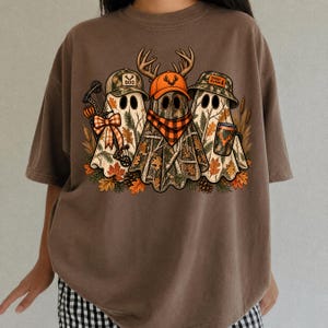 May include: A brown t-shirt featuring a graphic of three ghosts wearing camouflage hats. The ghosts are decorated with hunting gear, including a bow, a plaid scarf, and a mug. The design includes autumn leaves and the text BOO and SPOOKY SEASON.