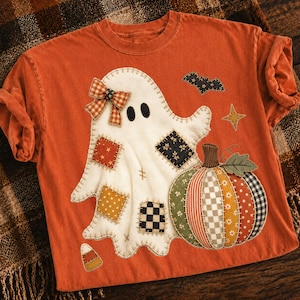 Op de afbeelding: Een roestoranje T-shirt met korte mouwen en een grote applicatie van een witte geest met een geruite strik, versierd met diverse stoffen lapjes. Naast de geest staan een veelkleurige patchwork pompoen, een kleine zwarte vleermuis, een ster en een snoepje.