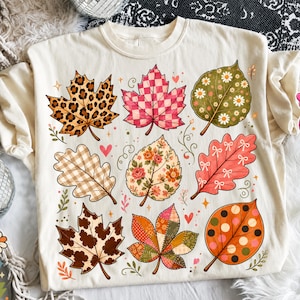 May include: A cream-colored t-shirt featuring a graphic print of nine stylized autumn leaves. Each leaf displays a unique pattern, including leopard print, pink and white checkers, floral designs, gingham, polka dots, and patchwork motifs.