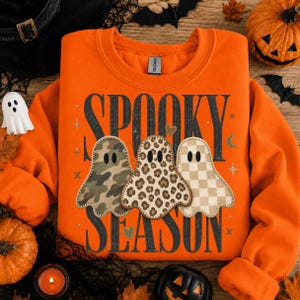 Peut inclure: Un sweat-shirt orange à col rond avec le texte noir SPOOKY SEASON. Le motif comprend trois fantômes avec des motifs différents : camouflage, imprimé léopard et damier beige, agrémentés de petites étoiles, de cœurs et de lunes.