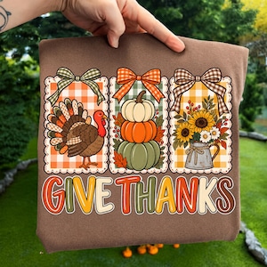 May include: A brown t-shirt featuring three rectangular panels with orange and white gingham backgrounds, each topped with a bow. The panels display a turkey, a stack of three pumpkins, and a pitcher of sunflowers. The text "GIVE THANKS" is printed below.