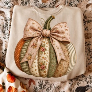 May include: A cream-colored crewneck sweatshirt featuring a large, central graphic of a pumpkin. The pumpkin is composed of various fabric-like segments in orange, green, and beige, decorated with floral, gingham, and paisley patterns, topped with a large bow.