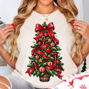 May include: A cream-colored t-shirt featuring a graphic of a decorated Christmas tree. The tree is adorned with red ribbons, a large red bow at the top, gold stars, red and white striped candy canes, and various red and white ornaments.