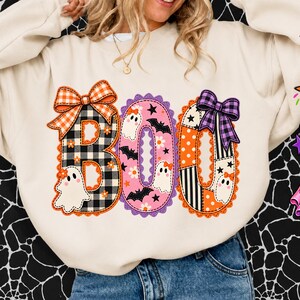Könnte beinhalten: Ein cremefarbenes Sweatshirt mit Rundhalsausschnitt und dem Wort BOO in großen, dekorativen Buchstaben. Das B ist schwarz-weiß kariert mit einer orangefarbenen Schleife, das O ist rosa mit Geistern und Fledermäusen, das zweite O ist orange-schwarz mit einer violetten Schleife.