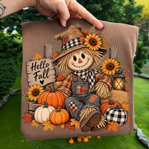 May include: A brown t-shirt featuring a graphic of a scarecrow wearing a hat and scarf, surrounded by sunflowers, pumpkins, and a lantern. A wooden sign with the text Hello Fall is visible on the left. The design includes autumn leaves and hay.