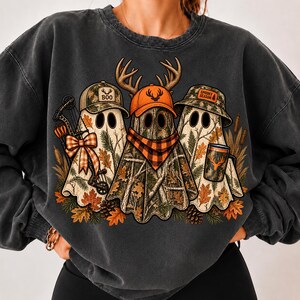 May include: A charcoal gray crewneck sweatshirt featuring a graphic of three ghosts. The left ghost wears a camo cap with the text BOO and holds a bow. The center ghost wears an orange cap and plaid bandana. The right ghost wears a camo bucket hat with the text SPOOKY SEASON and holds a mug with a deer silhouette.