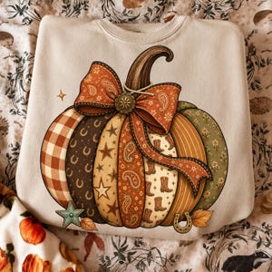 May include: A cream-colored crewneck sweatshirt featuring a large graphic of a pumpkin composed of vertical panels with various patterns including gingham, horseshoes, stars, paisley, and cowboy boots, topped with a large orange paisley bow.