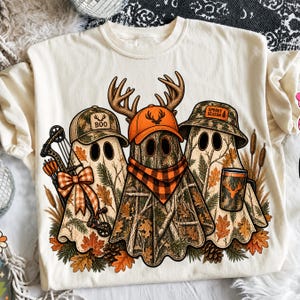 May include: A cream-colored t-shirt featuring a graphic of three ghosts wearing camouflage hats. The ghosts are decorated with orange plaid accents, a compound bow, and a mug with a deer silhouette. The text reads "BOO" and "SPOOKY SEASON".