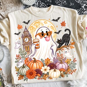 May include: A cream-colored t-shirt featuring a graphic of a ghost wearing orange sunglasses and blowing a bubble, holding a coffee cup. The design includes a clock tower, a black cat, pumpkins, autumn leaves, and small ghosts on a moonlit background.