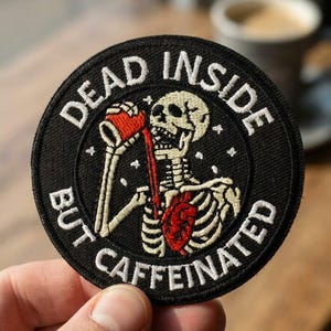 May include: A circular black embroidered patch featuring a white skeleton drinking from a red cup. A red anatomical heart is visible on the skeleton's chest. The text "DEAD INSIDE BUT CAFFEINATED" is embroidered in white around the border.
