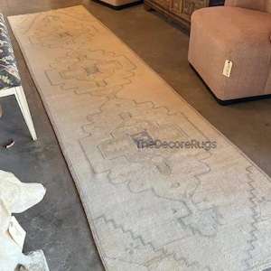 May include: A long, rectangular runner rug in a light beige tone featuring a repeating geometric pattern in muted blue and grey outlines. The text TheDecoreRugs is printed in the center of the rug. The rug has a flat, woven texture throughout.