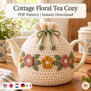 Op de afbeelding: Een crèmekleurige gehaakte theemuts met een trekkoord aan de bovenkant met groene linten en een decoratieve band van roze en gele bloemen met groene bladeren. De tekst Cottage Floral Tea Cozy PDF Pattern Instant Download is zichtbaar.