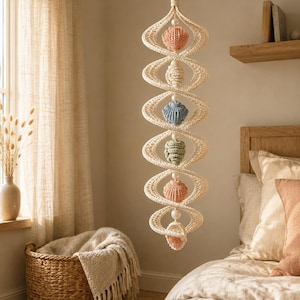 May include: A vertical macrame wall hanging featuring a cream-colored spiral structure. Interspersed within the spiral are six crocheted shell-shaped ornaments in shades of peach, cream, blue, sage green, and light pink, accented by small white beads.