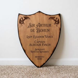 May include: A shield-shaped wooden plaque with a dark burnt edge, featuring two engraved dragons at the top corners and the text: Sir Arthur de Bohun, Lady Eleanor Vance, Captain Alistair Finch, Sergeant Rowena Kendrick, and Memento Mori.