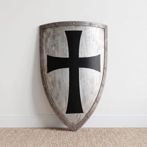 May include: A heater-style shield featuring a weathered, metallic silver finish and a prominent, bold black cross centred on its surface. The shield is bordered by a raised metal rim secured with evenly spaced rivets along the entire perimeter.