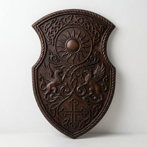 May include: A dark brown wooden shield featuring intricate carvings of two winged griffins facing each other, a central sun motif, a cross at the bottom, and decorative Celtic knotwork borders with a rope-like edge design throughout the piece.