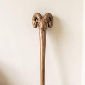 May include: A hand-carved wooden walking stick featuring a detailed ram head at the top. The sculpture displays the natural brown grain of the wood, with prominent curved horns and facial features of the ram integrated into the handle design.