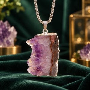 Peut inclure: Un collier pendentif composé d'une tranche de cristal d'améthyste brute et dentelée aux structures cristallines violettes et blanches, bordée d'une finition argentée métallique, suspendu à une chaîne à maillons carrés en acier inoxydable.