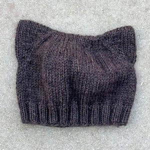 Puede incluir: Un gorro de punto color marrón oscuro que presenta dos formas puntiagudas en la parte superior que recuerdan a orejas. El gorro está confeccionado con hilo grueso en punto jersey y rematado con un borde de punto elástico acanalado.