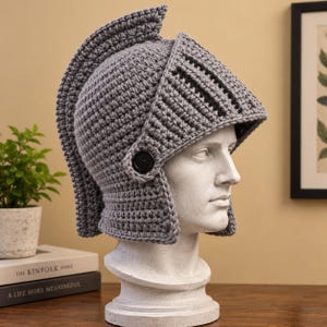 Puede incluir: Un casco de ganchillo gris con una cresta prominente y una visera, diseñado como un casco de caballero clásico. El casco presenta una construcción de hilo texturizado, un botón negro único en el lateral y una forma estructurada que cubre la cabeza.